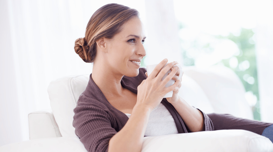 Pre-overgang klachten herkennen: Een vrouw zit met een kop koffie of thee op de bank, mogelijk nadenkend over veranderingen in haar lichaam.